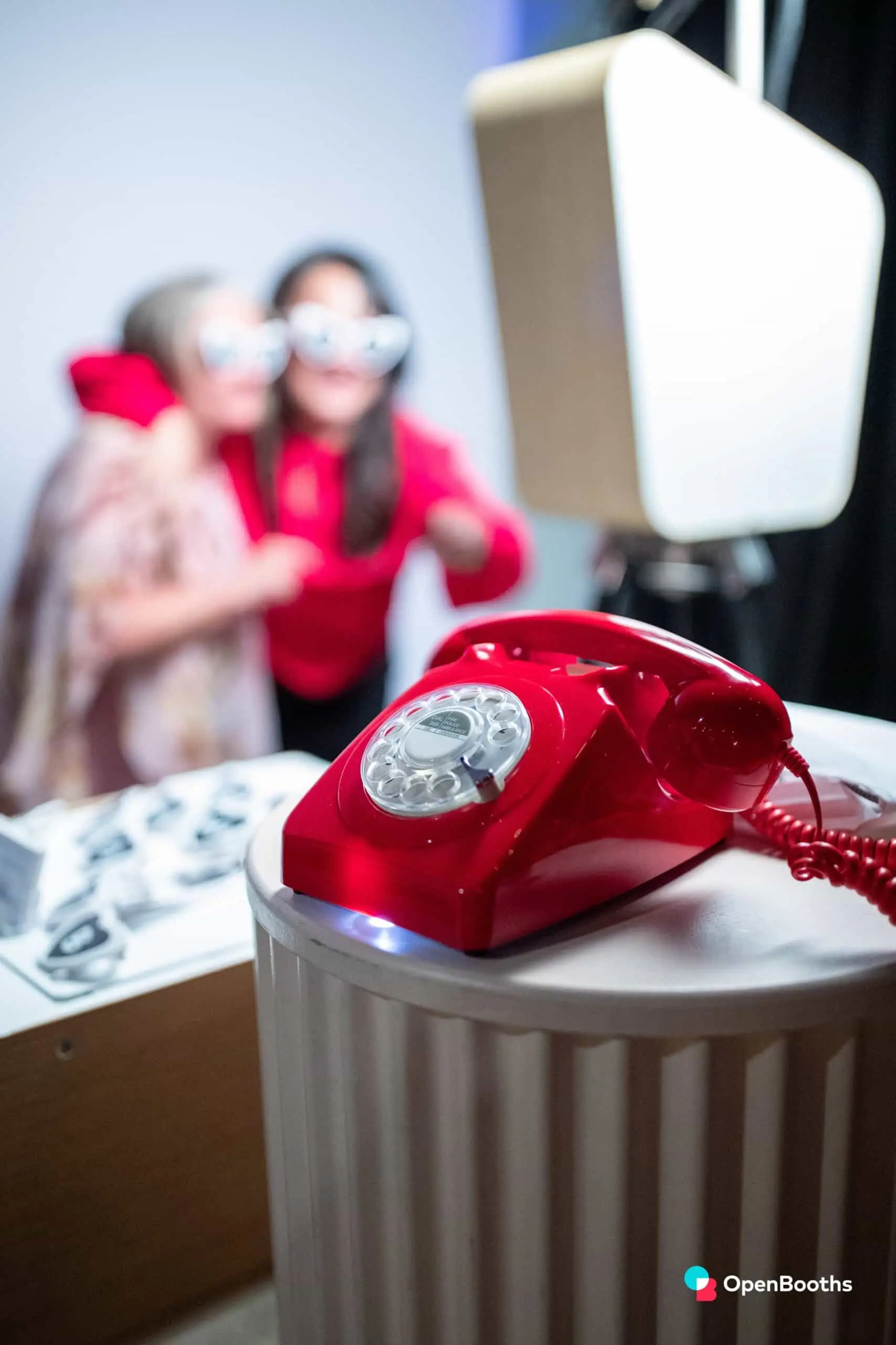 Telefon roșu cu disc pe un piedestal alb, cu invitați în fundal