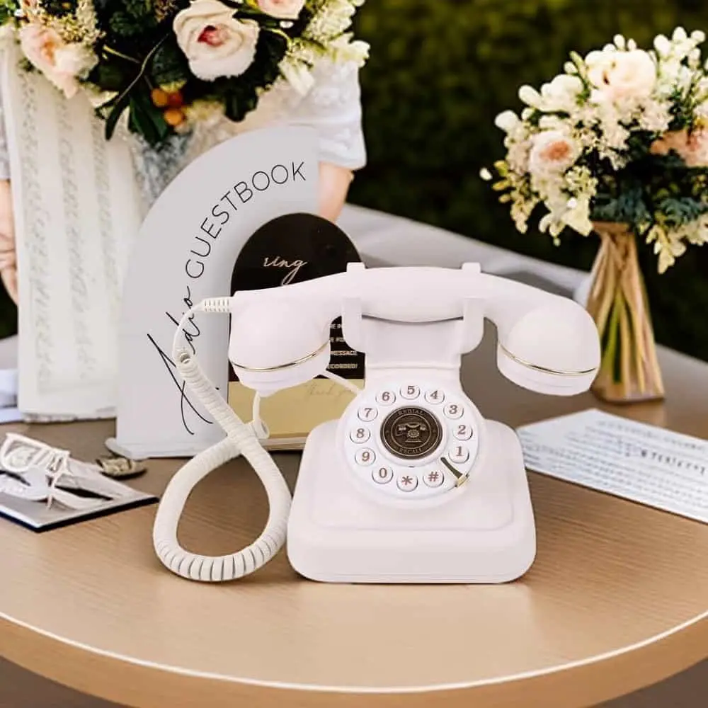 Telefon retro alb Elegance70 White pe o masă rotundă, lângă un panou guestbook și flori