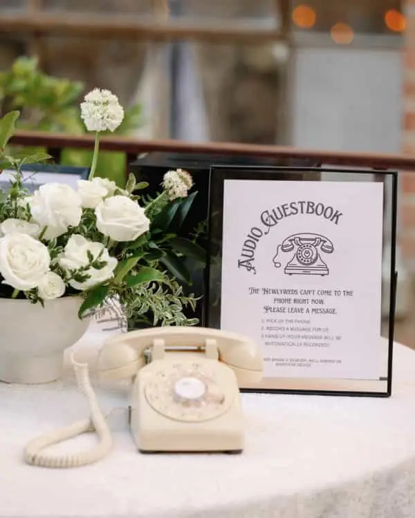 Telefon crem cu disc pe o masă rotundă, lângă un panou Audio Guestbook și trandafiri albi